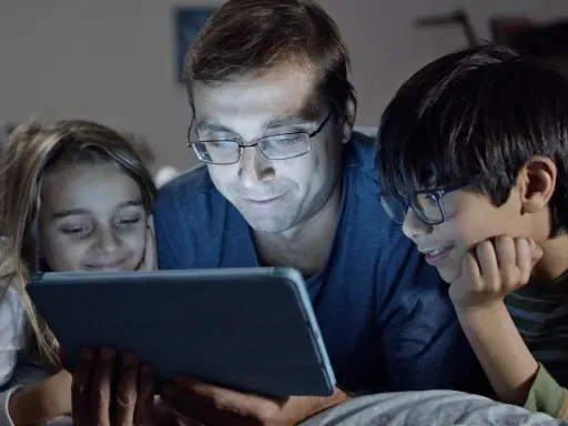 Padre y dos hijos ven una tablet, sonriendo. Podrían estar aprendiendo sobre los riesgos de las redes sociales o disfrutando de contenido familiar.