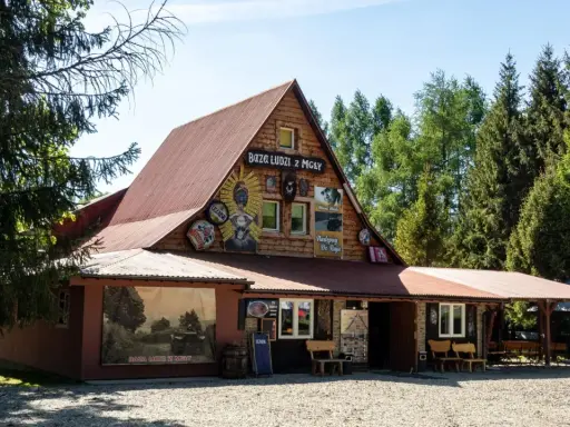 Drewniany budynek z napisem "Baza Ludzi z Mgły" i malowidłami. Obok duży mural z krajobrazem i napisem "Baza Ludzi z Mgły".