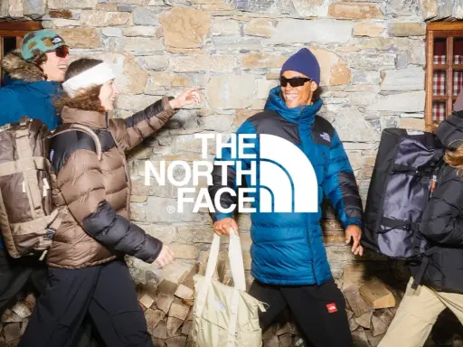 Cztery osoby w kurtkach The North Face, z plecakami i torbami, na tle kamiennej ściany. Jak rozpoznać podróbkę? Sprawdź detale!