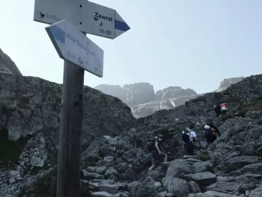 Grupa turystów wspina się po kamienistym szlaku w Tatrach. Tabliczka wskazuje kierunek na Zawrat.