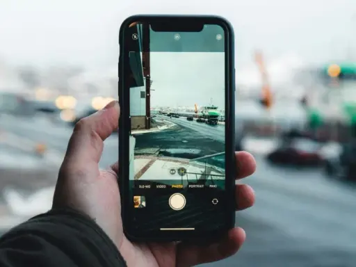 Dłoń trzyma aparat na telefon, rejestrując zimowy widok portu z zielonym statkiem i samochodami na drodze.