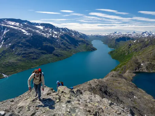 Vandrare klättrar på en bergskam med en turkos sjö nedanför. Besseggen svårighetsgrad är utmanande men belönande.