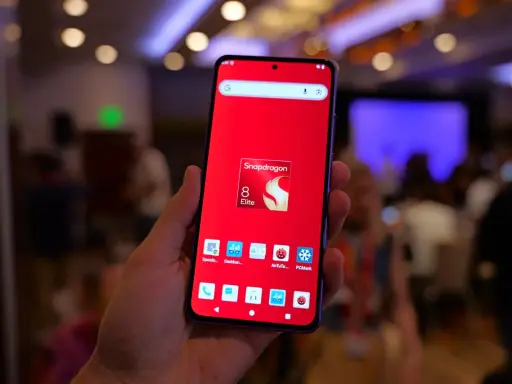 Dłoń trzyma smartfon z czerwonym tłem i ikoną Snapdragon 8 Elite. Widoczne ikony benchmarków, sugerujące testowanie wydajności procesora.