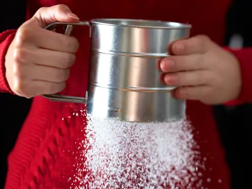 Hände halten einen Siebbecher, aus dem feiner Zucker rieselt. So einfach lässt sich puderzucker selber machen für süße Kreationen.