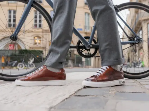 Eleganckie sneakersy do garnituru w brązowym kolorze, zestawione z szarymi spodniami, na tle miejskiego krajobrazu z rowerem.
