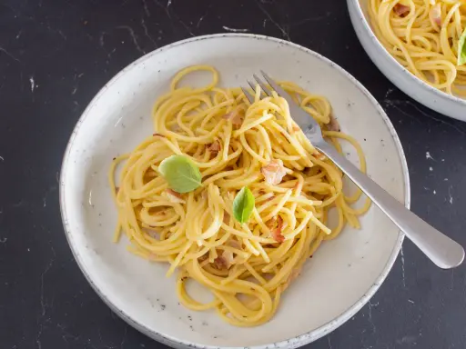 Ein Teller mit cremiger Spaghetti Carbonara, garniert mit Basilikum und Speckstückchen.