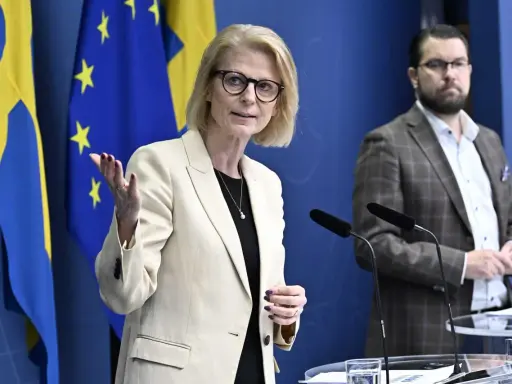 Kvinna med glasögon talar vid presskonferens med svenska och EU-flaggor bakom. Viktig information om folkräkning Sverige.