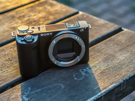 Czarny aparat Sony Alpha ZV-E10 bez obiektywu leży na drewnianym stole, oświetlony ciepłym światłem zachodzącego słońca.