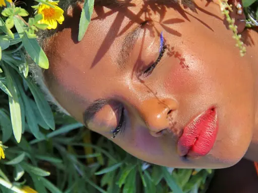 Un volto immerso tra fiori, con un trucco festival audace: eyeliner blu elettrico e labbra rosse vibranti.