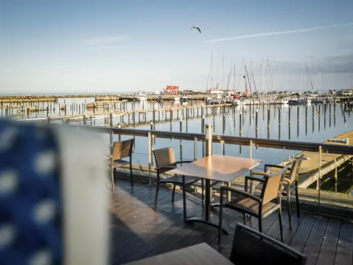 Ein Tisch mit Stühlen auf einer Holzterrasse mit Blick auf einen Yachthafen. Segelboote liegen im Wasser, die Sonne wirft lange Schatten. Ein Möwe fliegt über den Hafen. Damp 2000.
