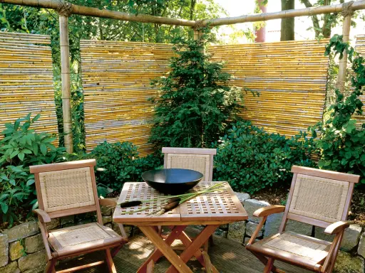 Ein lauschiger Sitzplatz mit einem selbstgemachten Zaun aus Bambus. Holzmöbel und üppige Pflanzen schaffen eine natürliche Atmosphäre.