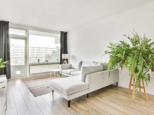 Soggiorno in casa stile nordico con divano grigio, tappeto a righe, piante verdi e grandi finestre.