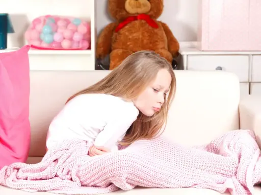 Bambina di 12 anni con mal di stomaco, rannicchiata sul divano sotto una coperta rosa.