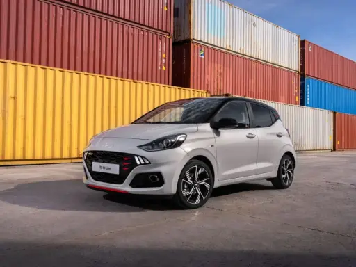 Szary Hyundai i10 N Line z czerwonymi akcentami na grillu i zderzaku, zaparkowany przed stosem kolorowych kontenerów. Sprawdź atrakcyjną hyundai i10 cena!