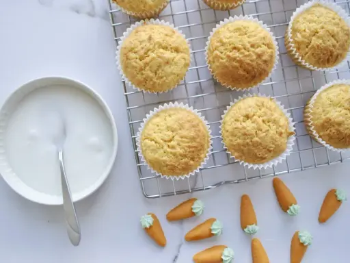 Goldbraune Karottenmuffins auf einem Gitter, daneben eine Schüssel mit Zuckerguss und süße Marzipankarotten. Perfekt für ein Karottenmuffins Rezept!