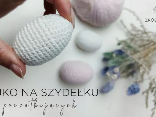 Ręcznie robione, białe jajko na szydełku, idealne na początek przygody z szydełkowaniem. W tle pastelowe włóczki i suszone kwiaty.