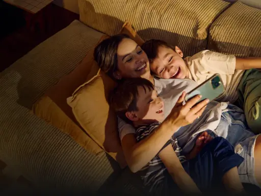 A mother and two sons laugh while looking at a smartphone on a couch.