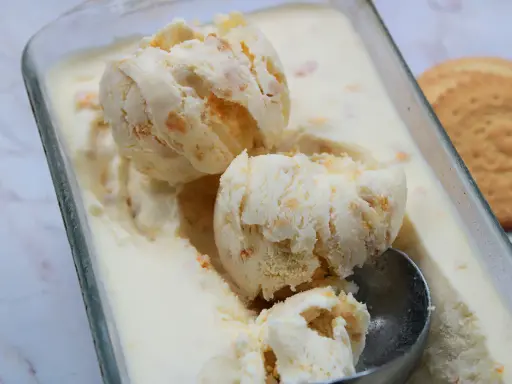 Pyszne lody ciasteczkowe w szklanym naczyniu, z widocznymi kawałkami ciastek i gałkami lodów. Obok leży okrągłe ciastko.