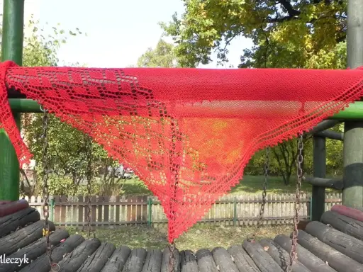 Czerwony, ażurowy szal, wykonany według darmowych wzorów z Ravelry, wisi na drewnianej konstrukcji placu zabaw.
