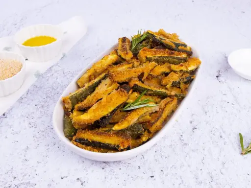 Zucchine gratinate al forno, dorate e croccanti, servite in un piatto ovale con rosmarino fresco. Accanto, ciotoline con pangrattato e olio.