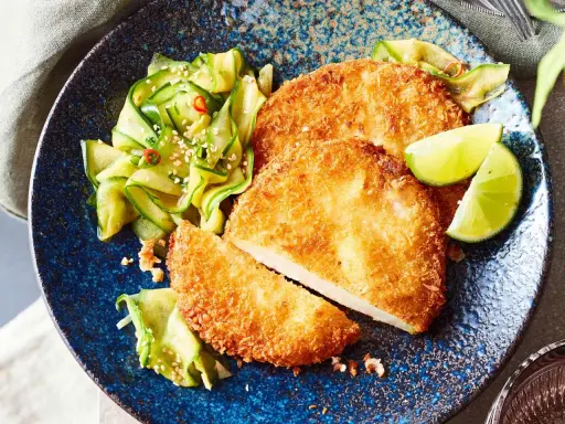 Knuspriges Kohlrabi-Schnitzel mit Gurkensalat und Limettenspalten auf blauem Teller.