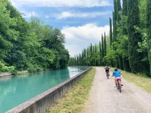 Bambini in bicicletta lungo la ciclovia del Mincio, con il canale turchese e filari di cipressi.