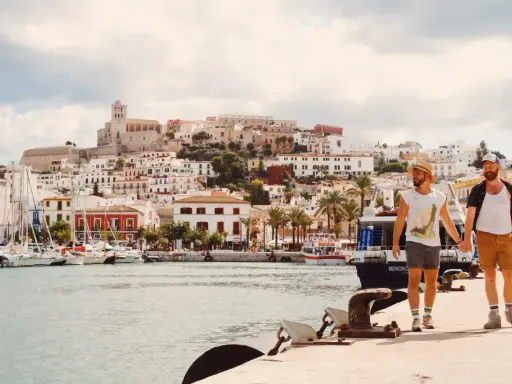 A gay couple walks hand-in-hand along a harbor in Spain, with a white-washed town and ancient fortress rising behind them.