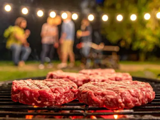 Frische Burger-Patties liegen auf dem Grill. Im Hintergrund feiern Freunde unter Lichterketten. So macht man Hackfleisch selber für den perfekten Grillabend.