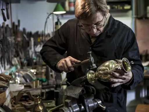 Un orfèvre au travail, martelant délicatement un vase en métal dans son atelier rempli d'outils.