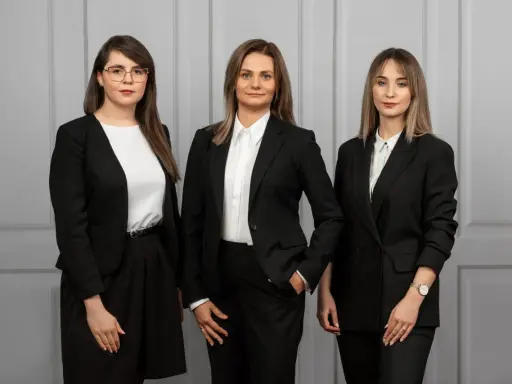 Trzy kobiety w czarnych garniturach stoją przed szarą ścianą.