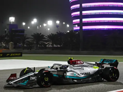 Silberner F1-Rennwagen mit Petronas-Sponsoren fährt bei Nacht auf der Rennstrecke in Bahrain. Flutlicht und ein beleuchtetes Gebäude im Hintergrund.