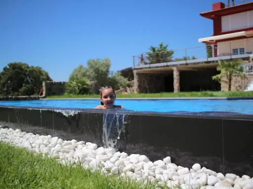 Niña sonriente en una piscina desbordante, con agua cayendo sobre piedras blancas y césped verde. Al fondo, una casa moderna y árboles frondosos bajo un cielo azul.