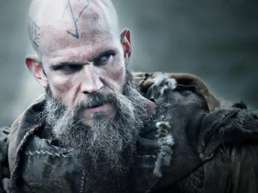 Uhtred z Bebbanburga, wojownik z tatuażami na głowie i długą, siwą brodą, spogląda groźnie.