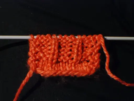 En orange stickad provlapp med ett mönster av vriden resår på en sticka. En lös tråd hänger ner.