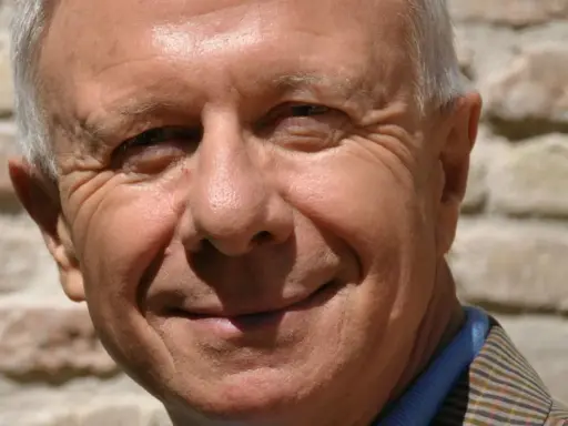Primo piano sorridente di padre Alberto Maggi, con rughe d'espressione e capelli grigi, sullo sfondo di un muro di mattoni.