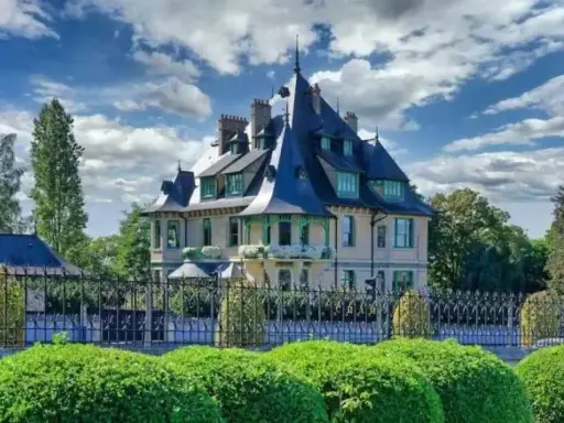 La villa Demoiselle, une demeure de caractère avec ses toits pointus et sa façade claire, se dresse fièrement derrière une grille ouvragée et des buissons taillés.