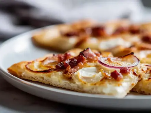 Tranches de flammekueche recette originale sur une assiette blanche, garnie de lardons, oignons rouges et fromage crémeux.