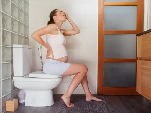 Ciężarna kobieta siedzi na toalecie, trzymając się za brzuch i czoło. W łazience widać bidet, co sugeruje lewatywa przed porodem.