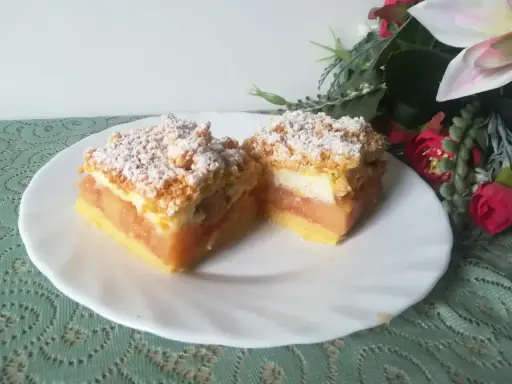 Pyszna szarlotka z bezą i kruszonką, z jabłkami i cukrem pudrem, na białym talerzu, obok kwiatów.