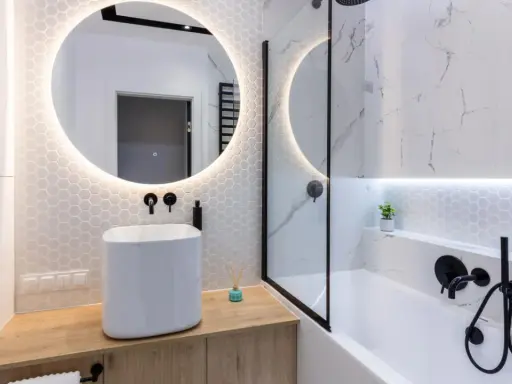 Elegante bagno senza finestra con lavabo bianco, specchio rotondo illuminato e doccia con finiture nere.