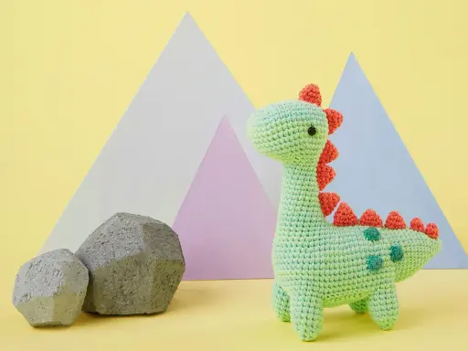 Adorable dinosaurio crochet verde con púas naranjas y detalles verdes. Dos rocas grises y formas geométricas de colores pastel al fondo.