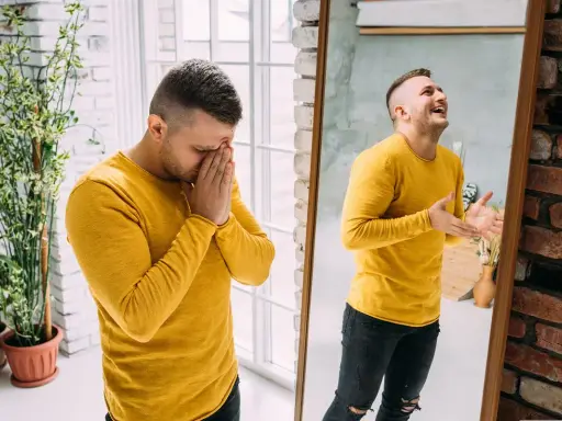 Mężczyzna w żółtej bluzce, raz ze smutkiem, raz z radością w odbiciu lustra. Obraz symbolizuje zmienne nastroje, jak u znanych Polaków z chorobą dwubiegunową.