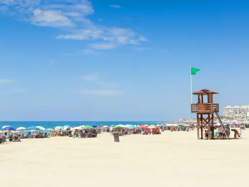 Sonniger Strand in Rota, Spanien, mit vielen bunten Sonnenschirmen und Menschen. Ein Rettungsschwimmerturm mit grüner Flagge steht am Meer.