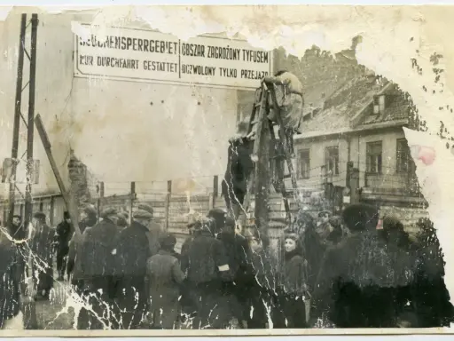 1941. Ludzie stoją przed znakiem "Obszar zagrożony tyfusem". W tle budynki i druty kolczaste.