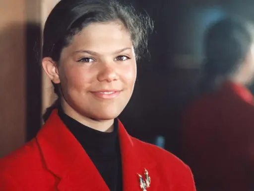 Kronprinsessan Victoria ung, med ett varmt leende, bär en röd kavaj och en svart polotröja. En vacker brosch pryder kavajen.