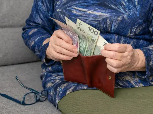 Dłoń starszej osoby wkłada pieniądze do portfela. Widać banknoty 100 zł. To wsparcie dla osób niepełnosprawnych, jak 500 plus dla niepełnosprawnych.