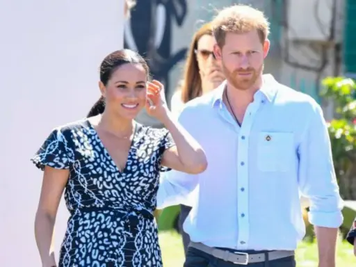 Meghan och Harry ler glatt under ett besök utomhus. Meghan bär en mönstrad klänning och Harry en ljusblå skjorta.