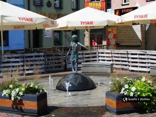 Fontanna z dziewczynką na kuli, otoczona donicami z kwiatami i parasolami. Urokliwe lubliniec atrakcje, idealne na spacer.