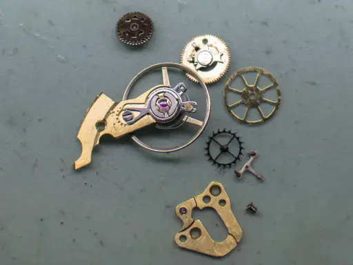 Parti di un orologio: ingranaggi, un bilanciere e altri componenti metallici disposti su una superficie grigia.