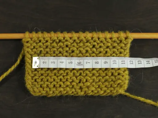 Knitted swatch with a measuring tape across it, showing stitches on wooden needles.